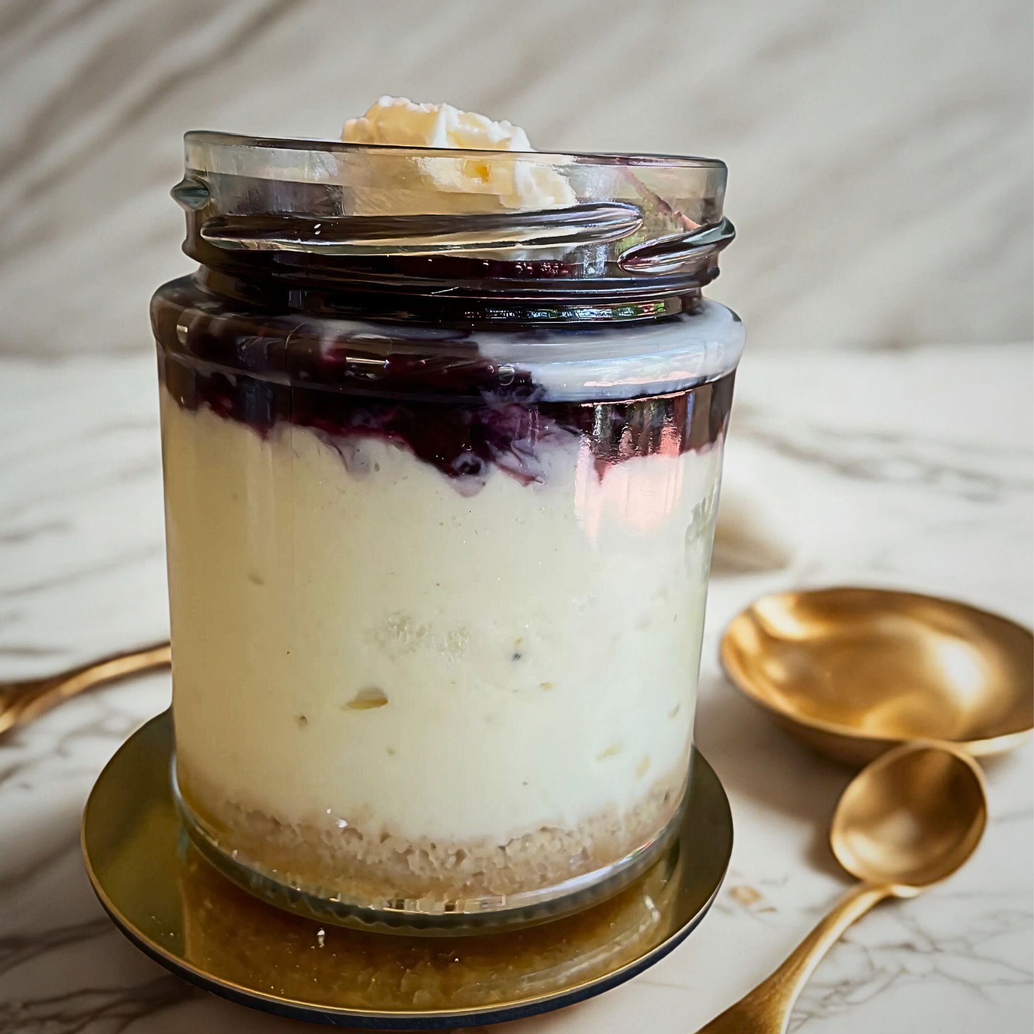 Blueberry cheesecake jar with layered blueberry compote on a gold plate, styled with gold cutlery on a marble surface.