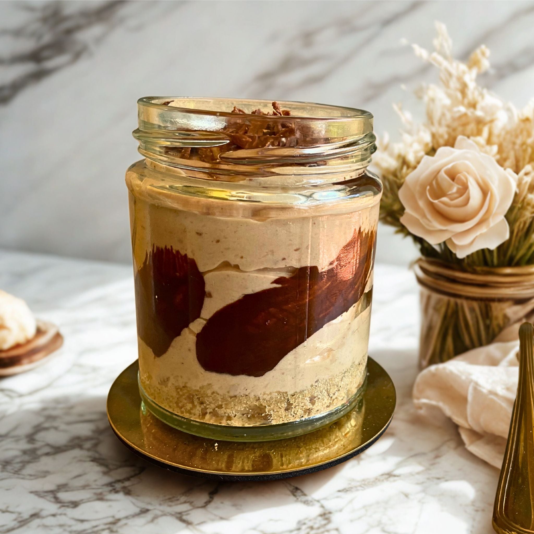 Front view of a Mocha Cheesecake jar from Seven Rivers, with rich coffee and chocolate layers, biscuit crust, and chocolate shavings, styled beside roses and a gold spoon.