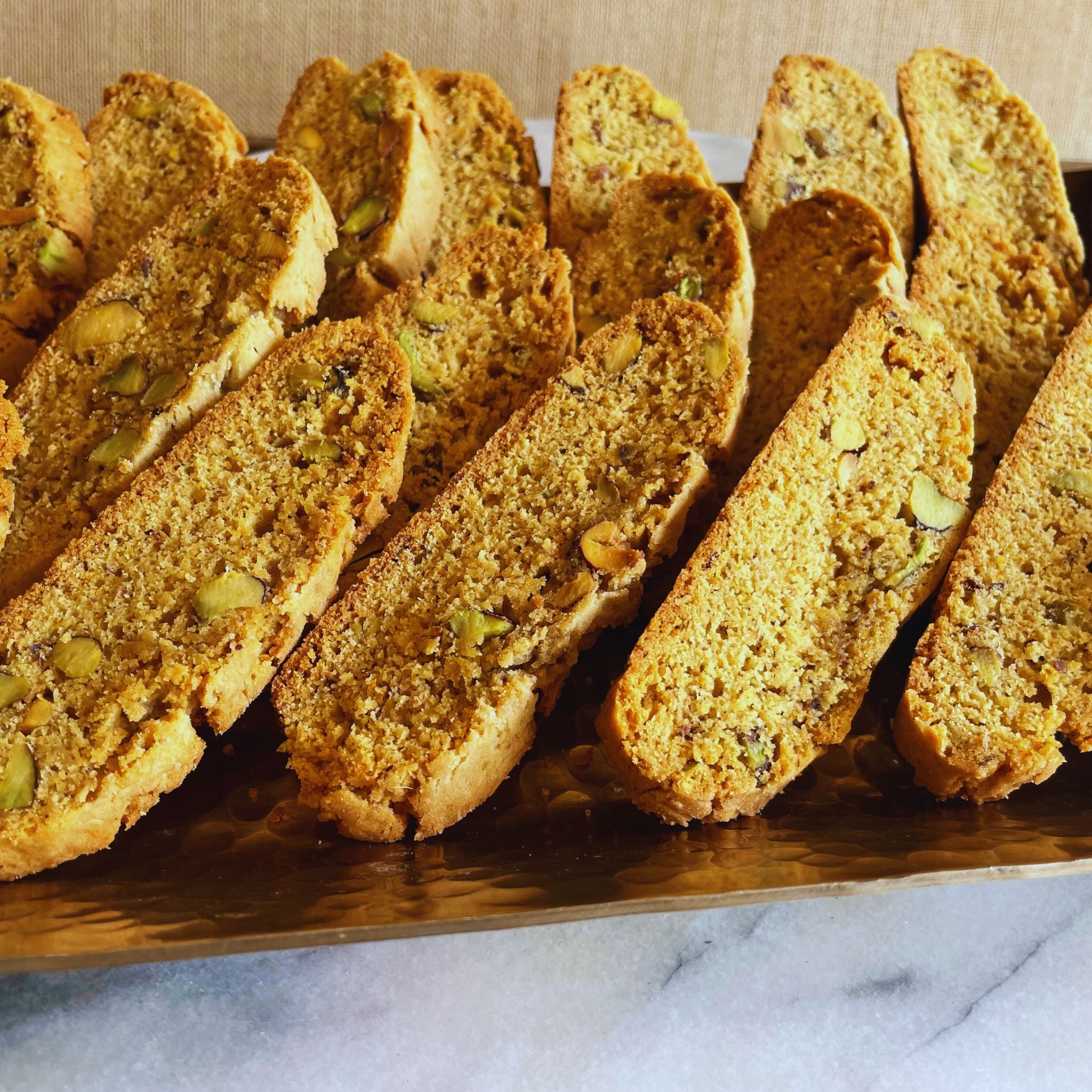 Crisp pistachio and orange biscotti lined up on a copper tray, highlighting their golden color and nutty texture.